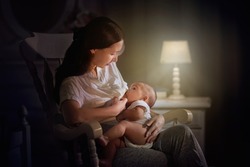 Mother and baby in dark bedroom. Asian mom with newborn boy in white rocking chair at night. Breastfeeding and infant care. Happy family at home. Bedtime and midnight feeding.