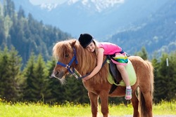 Kids riding pony in the Alps mountains. Family spring vacation on horse ranch in Austria, Tirol. Children ride horses. Kid taking care of animal. Child and pet. Little girl in blooming meadow.