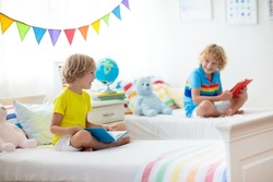 Child reading book in bed. Bedroom for two kids. Little boys read books on white bed with colorful bedding. Children nursery. Kid boy siblings play indoors. Nap and sleep time. Student doing homework.