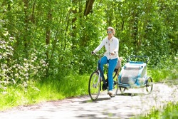 Young mother riding bicycle with baby bike trailer in sunny summer park. Fit active woman cycling with child. Safe transportation of little kids. Mom and children riding bikes. Family outdoor activity
