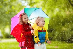 Little boy and girl play in rainy summer park. Children with colorful rainbow umbrella, waterproof jacket and coat playing in the rain. Kids walk in autumn shower. Outdoor fun by any weather. 
