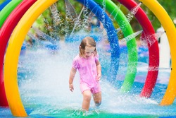 Happy little toddler girl running through a fountain having fun with water splashes in a swimming pool enjoying day trip to an aqua amusement park during summer family vacation