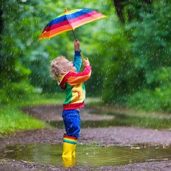 Little boy playing in rainy summer park. Child with colorful rainbow umbrella, waterproof coat and boots jumping in puddle and mud in the rain. Kid walking in autumn shower Outdoor fun by any weather

