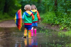 Little boy and girl play in rainy summer park. Children with colorful rainbow umbrella, waterproof boots jump in puddle and mud in the rain. Kids walk in autumn shower. Outdoor fun by any weather