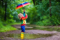 Little boy playing in rainy summer park. Child with colorful rainbow umbrella, waterproof coat and boots jumping in puddle and mud in rain. Kid walking in autumn shower. Outdoor fun by any weather

