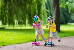 Children learn to ride scooter in a park on sunny summer day. Preschooler boy and girl in safety helmet riding a roller. Kids play outdoors with scooters. Active leisure and outdoor sport for child.