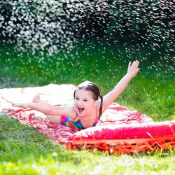 Little child playing with garden water slide. Kid jumping and splashing with gardening hose. Outdoor summer fun with backyard sprinkler for kids on hot sunny day.