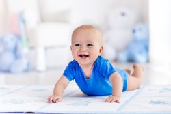 Adorable baby boy learning to crawl and playing with colorful toy in white sunny bedroom. Cute laughing child crawling on a play mat. Nursery interior, clothing and toys for little kids 