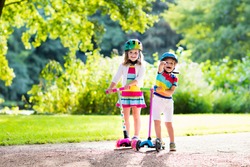 Children learn to ride scooter in a park on sunny summer day. Preschooler boy and girl in safety helmet riding a roller. Kids play outdoors with scooters. Active leisure and outdoor sport for child