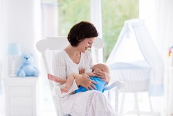 Mother and baby at home. Breastfeeding and healthy infant nutrition. Young woman nursing her newborn child sitting in white rocking chair in sunny nursery with crib and window. Parent and kid at home.