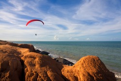 A para-glide soars over a tropical beach backed by red hills