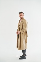 Full length portrait of young trendy man in beige trench coat over white studio background