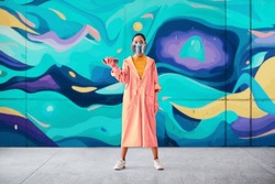 Full length portrait of graffiti woman painter in respirator mask standing near the wall with her paintings looking to camera. Street art concept