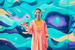 Portrait of female street artist in respirator mask standing near the graffiti wall with her paintings looking to camera. Urban art concept
