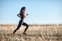 Asian girl in sportswear running across field, morning workout. Eastern girl doing sports. Side view of active athletic young woman running on blue sky background. Photo with copy space