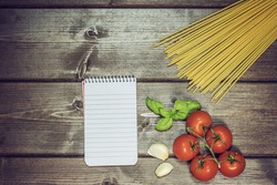 Blank notebook with lined paper, garlic cloves, basil leaves, spaghetti and tomatoes are lying on the wooden table. Edited as a vintage photo with dark edges.