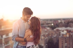 Couple in love standing and hugging on a building rooftop at sunset with cityscape in the background. Focus on the girl