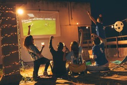 Group of young friends watching a football match on a building rooftop, drinking beer and cheering. Selective focus on the people in the middle