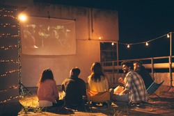 Group of young friends watching a movie on a building rooftop terrace, eating popcorn, drinking beer and having fun