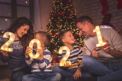 Parents celebrating New Years Eve at home with kids, sitting by the Christmas tree, holding illuminative numbers 2021 representing the upcoming New Year