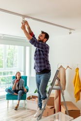 Couple in love moving in together, man standing on the ladder and changing the light bulb while woman is drinking coffee and relaxing