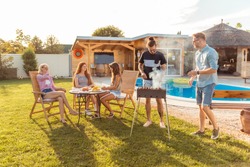 Group of young friends having a backyard barbecue party, grilling meat and having fun while spending sunny summer day outdoor by the swimming pool