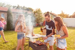 Group of cheerful young friends having a backyard barbecue party, grilling meat, drinking beer, playing badminton and relaxing on a sunny summer day outdoors