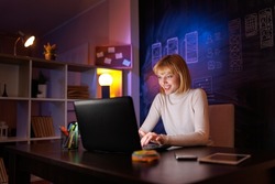 Woman sitting at her desk in home office working late at night using laptop computer; female web designer working overtime remotely from home