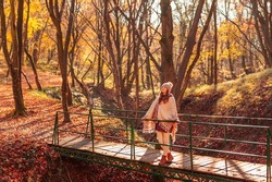 Woman walking down the bridge over a stream on the forest path covered with colorful fallen leaves, enjoying spending sunny autumn day outdoors in nature