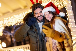 Beautiful young couple in love having video call with friends using smart phone while celebrating New Year in the city streets with Christmas lights all around