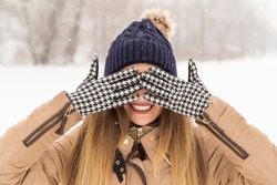 Beautiful woman wearing a warm winter clothes, hiding her eyes with hands in gloves, enjoying a snowy winter day in nature