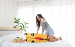 Young Asian woman trying to pack her clothes in to yellow suitcase preparing for travel summer trip on bed. Travel concept