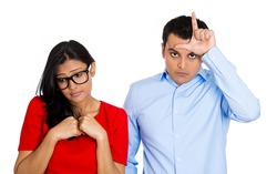 Closeup portrait of couple. Bully husband, man standing  upfront angry, loser sign on head, shy wife, nerdy woman wearing glasses looking downwards, isolated on white background. Human emotion culture