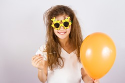Closeup portrait happy, smiling, funny looking little girl with sunglasses, holding orange balloon and needle about to burst bubble, isolated background. Human face expressions, emotions, feelings