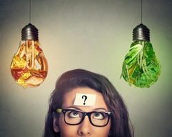 Woman in glasses question mark on head thinking looking up at junk food and green vegetables shaped as light bulb isolated on gray background. Diet choice right nutrition healthy lifestyle concept  