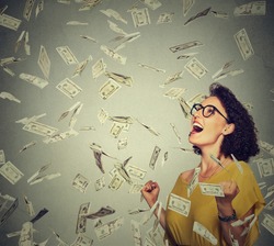 Portrait happy woman in glasses exults pumping fists ecstatic celebrates success under a money rain falling down dollar bills banknotes isolated on gray wall background with copy space  