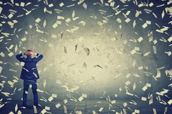 Rear back view of business man standing in front of a wall under money rain dollar banknotes falling down, hands on head wondering what to do next. Full body length of businessman facing the wall