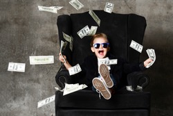 Happy shouting self-confident rich kid boy millionaire sitting with legs crossed in luxury armchair and throwing money dollars cash. Overspend.