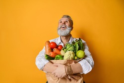 Cool old mature senior man with gray beard shopping hold grocery shopping bag with healthy organic vegetables looking up on yellow background