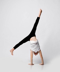 Slim athletic blonde kid girl is doing gymnastic acrobatics exercise at home in studio, handstand and stretching with her legs up on white background