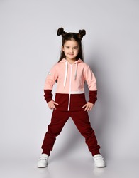 Frolic smiling kid girl with straight brunette hair with buns in two-colored pink brown sport suit hoodie and pants poses with legs wide apart and hands on hips