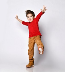 Little brunet kid in red jumper, brown pants and sneakers. He smiling, raised his hands and leg up, posing isolated on white studio background. Childhood, fashion, advertising. Full length, copy space