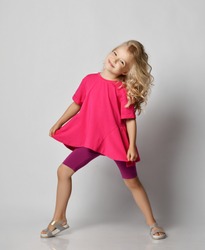 Beautiful positive little blonde girl with curly hair in stylish casual shorts and t-shirt posing and smiling over grey wall background. Fashion for children concept
