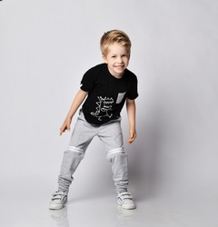 Playful frolic blond kid boy in sunglasses, black t-shirt with dinosaur print and gray pants stands leaning forwards going to run out playing catch-up over gray background