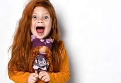 Little five-year-old red-haired girl happy screaming yelling. Child kid holds a red hair doll in her hands in an orange sweater on white background