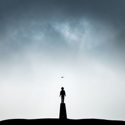 A lone boy standing in silhouette on a trig point beneath brooding clouds with a seagull flying overhead