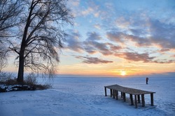 winter scene with a tranquil frozen lake. As the sun sets, the sky transforms into a masterpiece of vibrant colors, casting a warm, orange glow over the icy landscape and a charming wooden pier