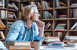 Dreamy thoughtful young teacher man male student hipster author glasses chin on hand study in library sit at desk write essay ready for exam do homework assignment smile look away education concept.