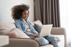 Young african ethnic woman remote worker student freelancer holding using laptop computer studying working at home online sitting on sofa. Serious black girl searching browsing internet in living room