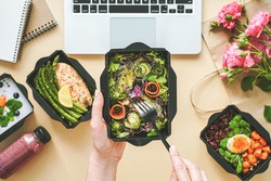 Female hands hold food delivery box having lunch at work from home office. Business woman worker eats salad take away nutrition daily healthy meal weight loss diet menu at workplace flat lay top view.